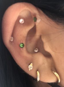 Ear of a brown-skinned person with gold hoop earrings an green and clear crystal d earseeds